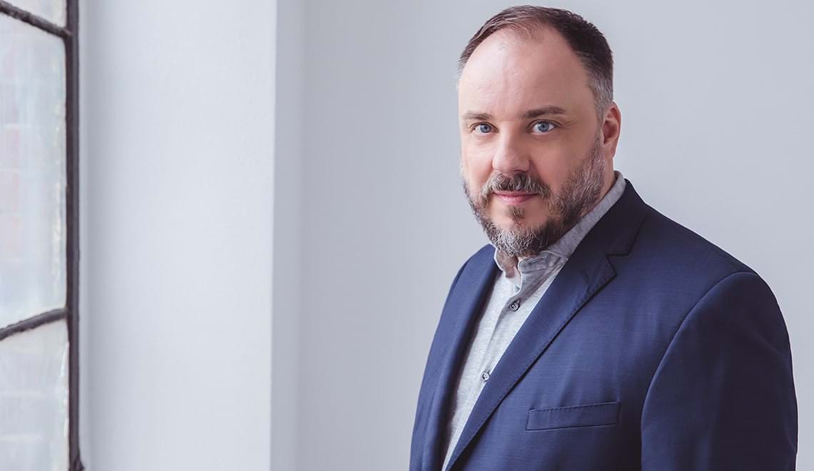 Matthias Goerne wears a blue suit jacket and a grey shirt. He is standing next to a window and in bright light, he has short dark hair, a beard and blue eyes.