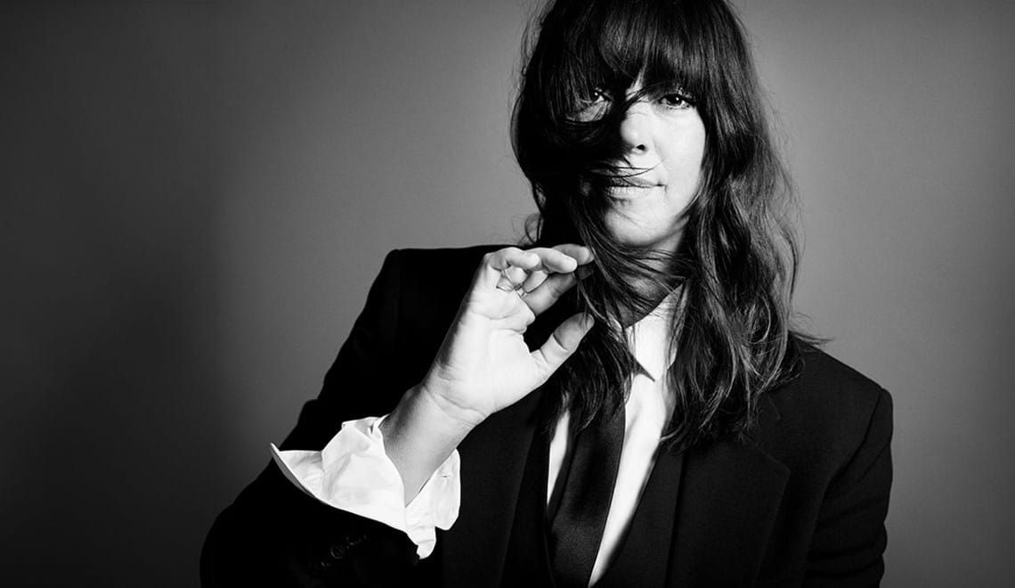 Cat Power wears a black suit and tie with a white shirt. They have long hair and a fringe and their hand is brushing their hair away from their face. The image is black and white.