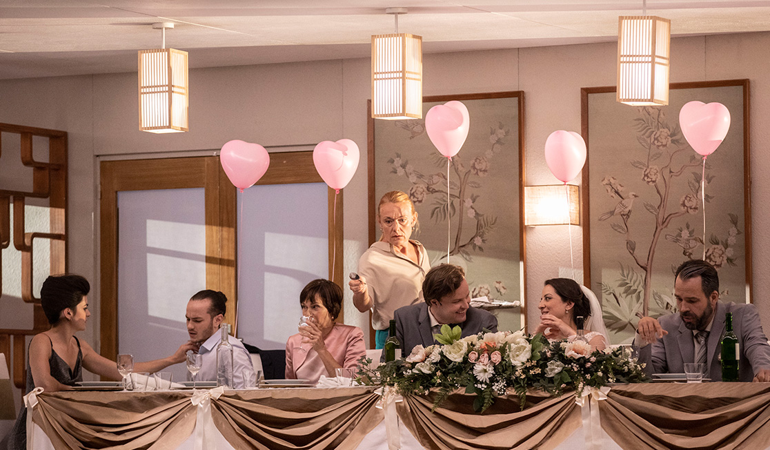 A long table with a bride and groom and wedding guests. There are pink heart balloons above them and someone is setting a spoon on the table with a serious expression.