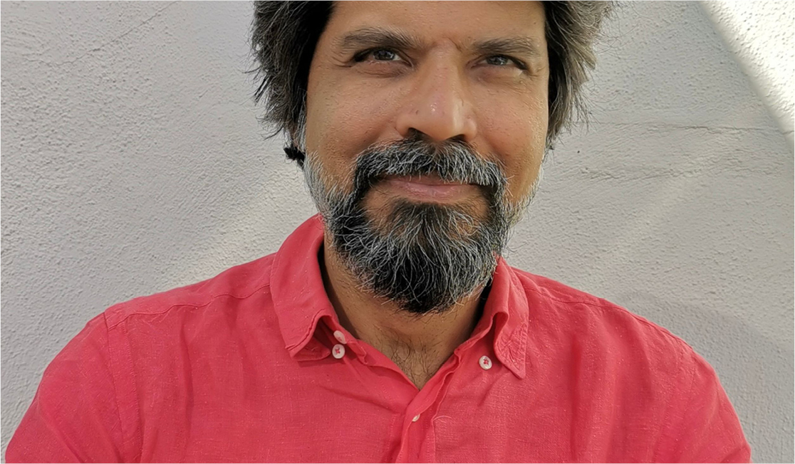 Pankaj Mishra wears a red shirt and smiles. He has a dark beard and short dark hair.