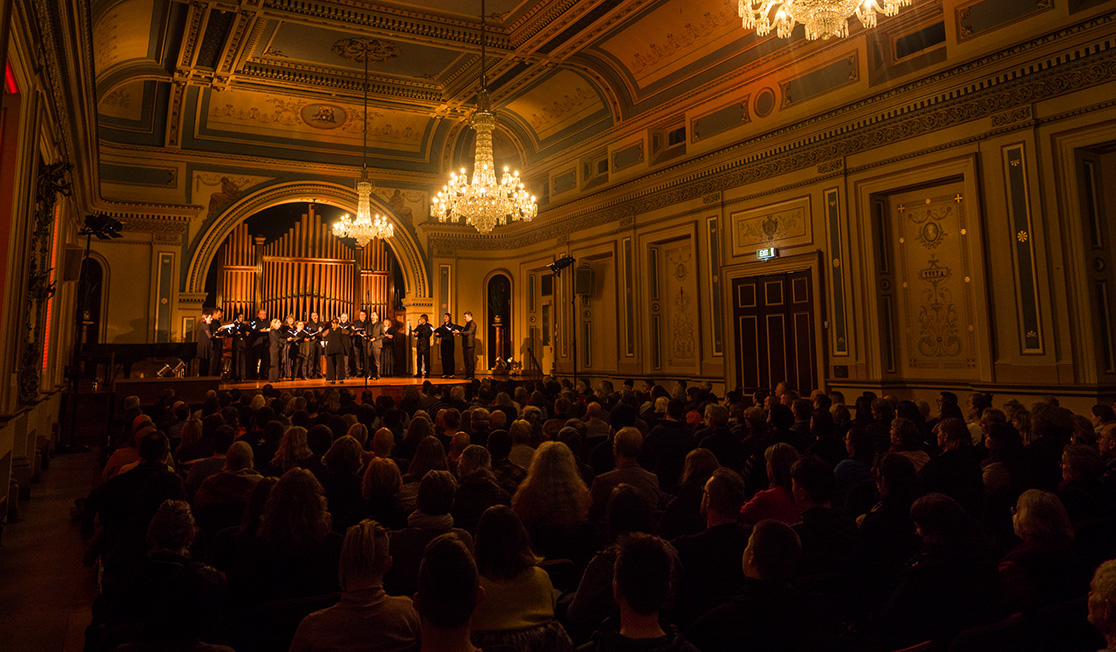 A group of singers dressed in black stands on a stage in a dimly lit hall. The heads of the audiences are visible with chandeliers overhead.